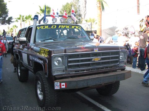 1990 Ford Bronco - brown Chevrolet Blazer prerunner marked Yolo Racing, with snorkel, roof lights and an American flag, sits parked among spectators.