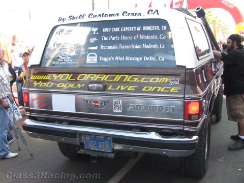 1990 Ford Bronco - rear of a brown Chevrolet SUV displays sponsor names and Yolo Racing You Only Live Once lettering across the tailgate and windows.