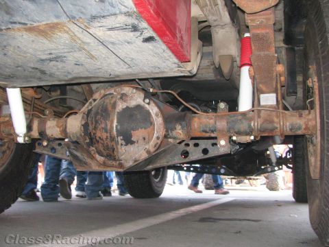 1990 Ford Bronco - under-vehicle view shows a red truck's rear differential, axle tubes and red-and-white shocks above a concrete floor.