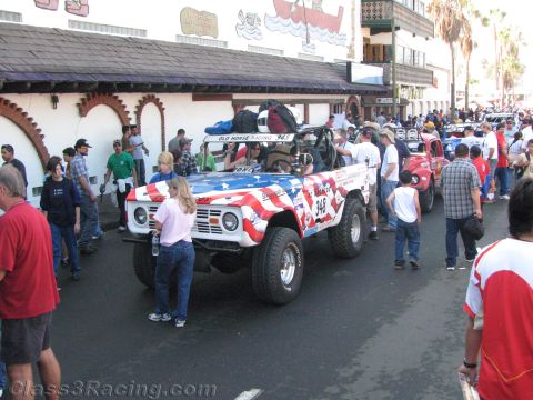 1990 Ford Bronco - vintage open-body off-road vehicle painted in American flag colors sits on a crowded street lined with spectators and palm trees.