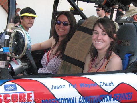 1990 Ford Bronco - Two smiling women sit in the cockpit of an open off-road buggy lettered with sponsor names at a racing event.