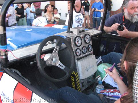 1990 Ford Bronco - cockpit of an open off-road buggy shows a chrome steering wheel and aluminum gauge panel while a bearded man photographs it.