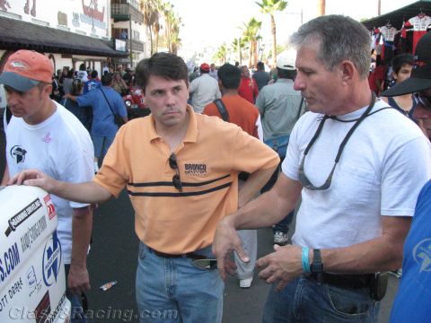 1990 Ford Bronco - Two men converse in a busy street, one wearing an orange striped polo, amid a crowd at an outdoor off-road event.