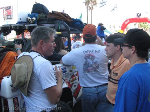 1990 Ford Bronco - group of men talk beside a race vehicle stacked with gear and spare tires in a crowded street under palm trees.