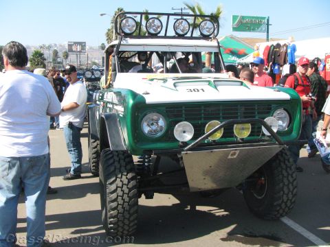 1990 Ford Bronco - green-and-white early Ford Bronco off-road race truck numbered 301 with a roof-mounted light bar parked in a crowd.