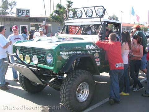 1990 Ford Bronco - green early Ford Bronco off-road racer numbered 301 with roof lights surrounded by spectators on a Mexican street.
