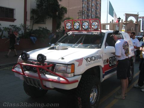 1990 Ford Bronco - white off-road race truck numbered 302 with Yokohama graphics and a roof light bar parked on a street.