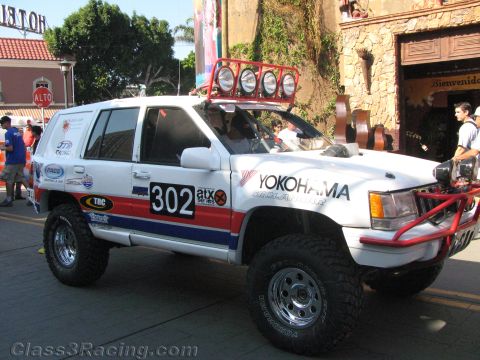 1990 Ford Bronco - Side view of a white SUV off-road racer numbered 302 with Yokohama and Land Rover service graphics and a roof light bar.