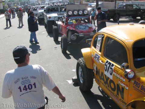 1990 Ford Bronco - yellow Baja Bug numbered 575 and other off-road vehicles staged on a street as spectators watch.