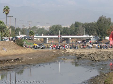 1990 Ford Bronco - Spectators line a dry, muddy riverbed near a concrete highway bridge where off-road vehicles gather.