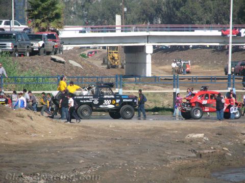 1990 Ford Bronco - black off-road truck numbered 300 and a red racer travel a road beside a riverbed under a concrete bridge.