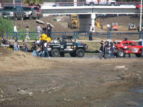 1990 Ford Bronco - black off-road truck numbered 300 and a red racer surrounded by spectators on a road beside a muddy riverbed.
