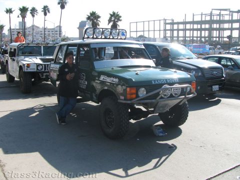 1990 Ford Bronco - green Range Rover off-road racer with a roof light bar and BFGoodrich graphics parked on a street with onlookers.