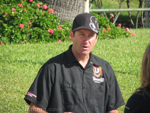 1990 Ford Bronco - man in a black cap and black team shirt speaks outdoors in front of pink flowering bushes and grass.