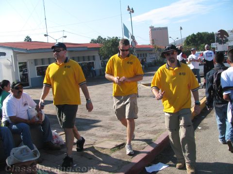 1990 Ford Bronco - Three men in matching yellow team polo shirts walk across a paved plaza as people look on.