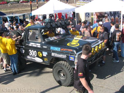 1990 Ford Bronco - black off-road race pickup numbered 300 with a roof-mounted light bar sits amid a crowd of spectators and crew at a desert racing event.