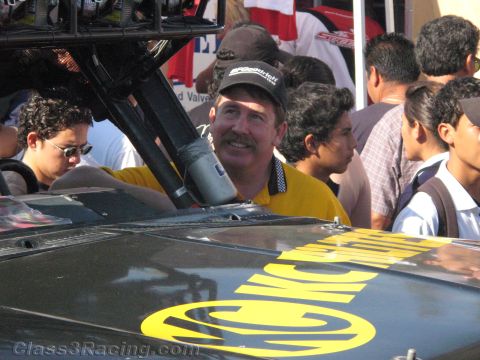 1990 Ford Bronco - smiling mustached man in a yellow shirt and dark cap stands beside a black race vehicle's roll cage amid a crowd of people.