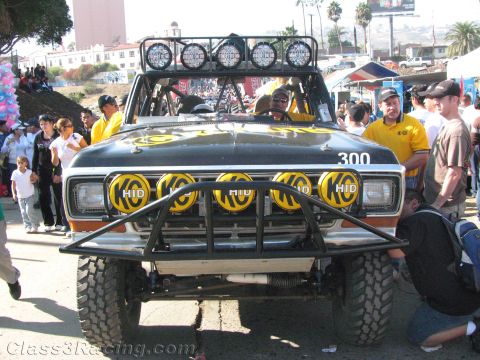 1990 Ford Bronco - Front view of a black off-road race truck numbered 300, fitted with a brush guard and five yellow HID driving lights on its roof bar, surro…