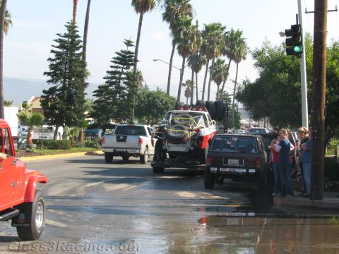 1990 Ford Bronco - off-road race buggy strapped to a trailer waits at a palm-lined street intersection with a green light while bystanders watch.