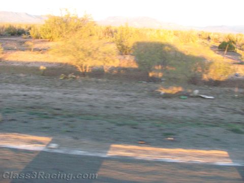 1990 Ford Bronco - motion-blurred view of desert scrubland and distant hills at golden hour, shot from a moving vehicle alongside a paved road edge.