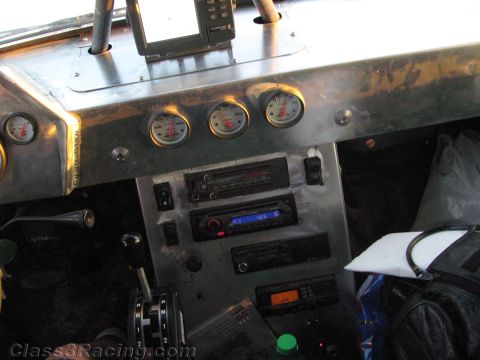 1990 Ford Bronco - bare-metal dashboard of a race vehicle showing three round white gauges, stacked aftermarket stereo and radio head units, and a shifter.