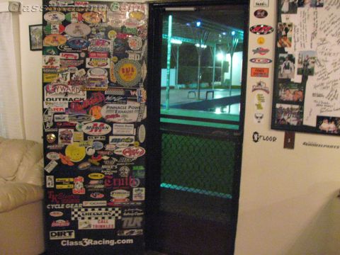 1990 Ford Bronco - interior room wall and doorway plastered with dozens of racing decals and stickers, beside a leather couch, with a lit pool visible through…