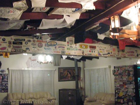 1990 Ford Bronco - dimly lit room with exposed beams draped in signed shirts and lined with stickers, set above pale leather couches and a curtained window.