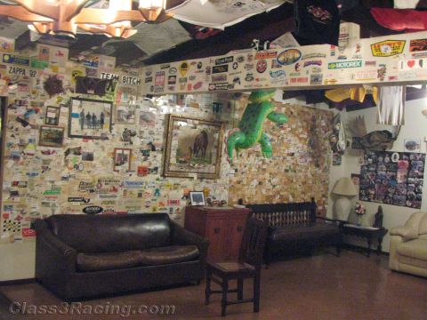 1990 Ford Bronco - lounge crowded with racing memorabilia: sticker-covered walls, framed pictures, a green lizard wall figure, and worn leather sofas around a…