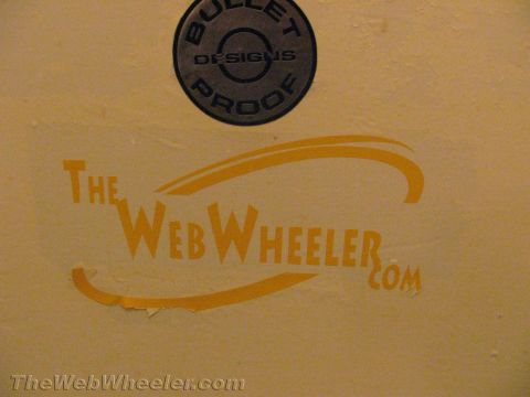 1990 Ford Bronco - Close-up of a yellow wall showing a round gray 'Bullet Proof Designs' decal above an orange 'TheWebWheeler.com' logo sticker.