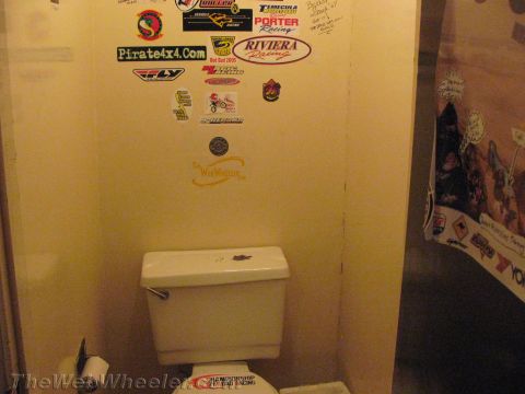 1990 Ford Bronco - small restroom with a white toilet beneath yellow walls plastered in racing stickers including Pirate4x4.com, Riviera Racing and TheWebWhee…