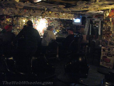 1990 Ford Bronco - dimly lit bar interior where several patrons sit at the counter, walls and ceiling plastered with stickers and racing memorabilia.