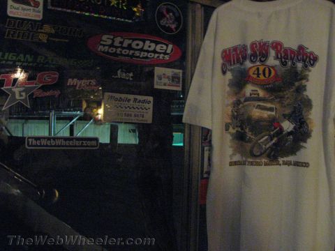 1990 Ford Bronco - white t-shirt printed with a 'Mike's Sky Rancho 40' off-road graphic hangs on a wall covered in racing stickers and signs.