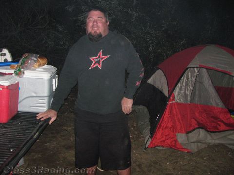 1990 Ford Bronco - smiling bearded man in a dark star-logo hoodie stands at a truck tailgate beside a cooler and a red dome tent at a nighttime campsite.