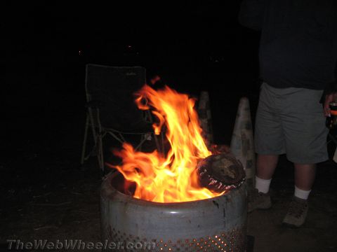 1990 Ford Bronco - bright campfire blazes inside a perforated metal drum at night, with a folding chair behind it and a person standing to the side.