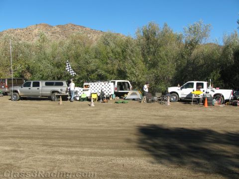 1990 Ford Bronco - desert staging area with pickup trucks, canopies, tents and a checkered flag lined up against trees beneath a rocky mountain.