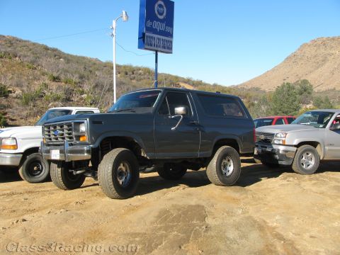 1990 Ford Bronco - lifted dark Ford Bronco on oversized tires is parked on dirt between other pickups beneath a blue roadside sign in hilly terrain.