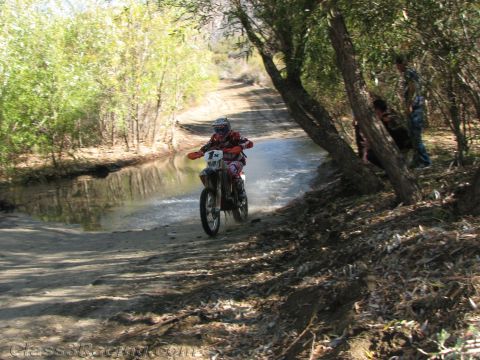 1990 Ford Bronco - motorcyclist in red riding gear powers a dirt bike through a shallow water crossing on a tree-lined dirt trail as spectators watch.