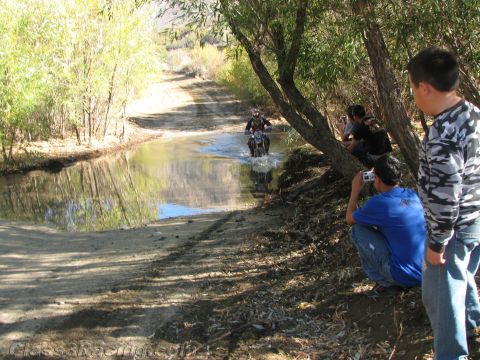 1990 Ford Bronco - Several spectators sit on a bank watching a dirt-bike rider splash through a shallow stream on a tree-lined dirt trail.