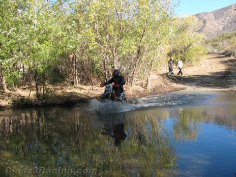 1990 Ford Bronco - dirt-bike rider sprays water while riding through a shallow stream toward the camera, with hills and trees in the background.