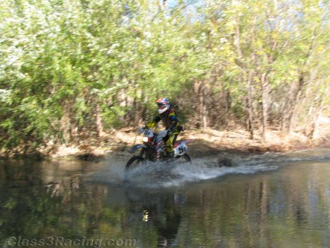 1990 Ford Bronco - dirt-bike rider in colorful gear throws up a spray of water while riding through a stream lined with green trees.