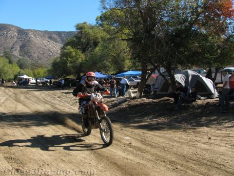 1990 Ford Bronco - dirt-bike rider kicks up dust along a wide dirt trail past campsite tents and parked vehicles beneath a rocky mountain.