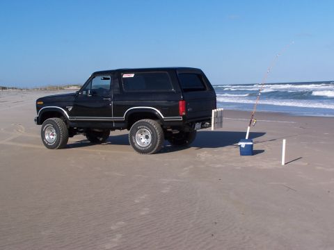 1983 Ford Bronco - A black 1983 Ford Bronco is parked on the beach next to a fishing rod and bucket.