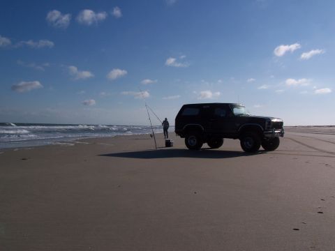 1983 Ford Bronco - A person fishes from the shore next to a black SUV on a sandy beach under a cloudy blue sky.