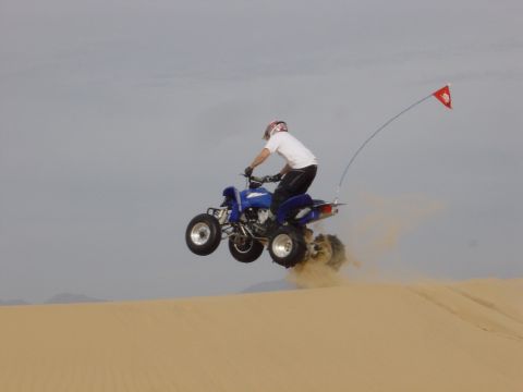 1986 Toyota Hilux - A rider on a blue ATV soars through the air over a sand dune, a red flag trailing behind.