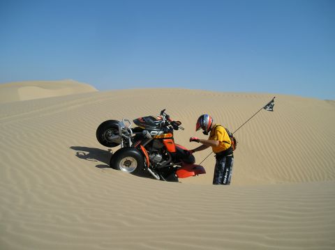 1986 Toyota Hilux - A person attempts to free an orange ATV that has become stuck in the sand dunes under a clear blue sky.