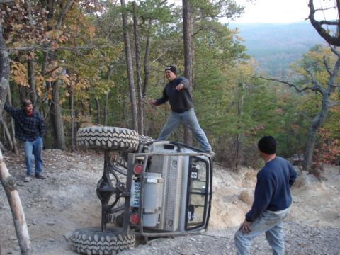 1978 Ford Bronco - An off-road vehicle has overturned on a steep, wooded trail, with onlookers observing the scene.