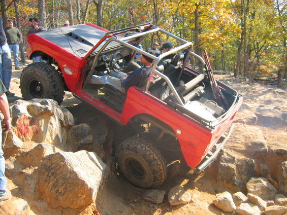 Red off-road vehicle with roll cage navigating rocky terrain. Hood has vents