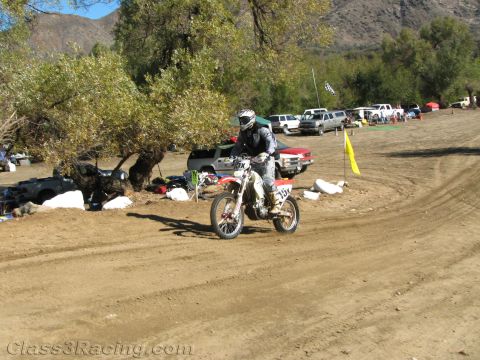 1990 Ford Bronco - dirt-bike rider leans through a banked dirt corner past parked pickups and a yellow course flag with hills behind.