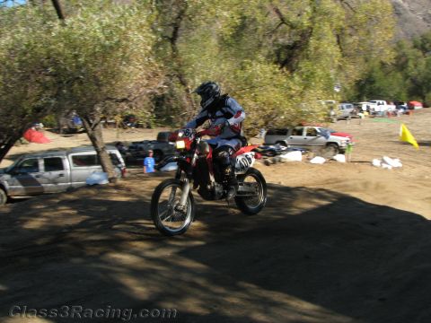 1990 Ford Bronco - rider on a red dirt bike catches air over a dirt rise with parked trucks, a course flag and trees in the background.