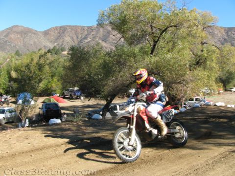 1990 Ford Bronco - dirt-bike rider in red gear rides a wide dirt trail past a campsite below a large rocky mountain ridge.